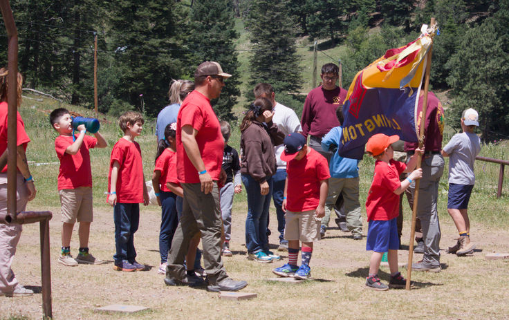 Cub Scouts and adult leaders gather outdoors at Wehinahpay Mountain Camp, with one Scout holding a colorful flag while others participate in a group activity surrounded by pine trees.