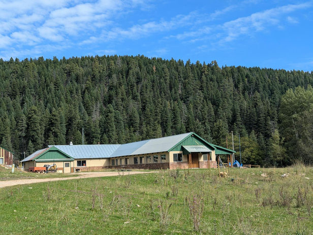 Wide exterior view of the Wehinahpay Mountain Camp dining hall and adjoining buildings, featuring green metal roofs and a grassy meadow, with densely forested mountains and a blue sky in the background.