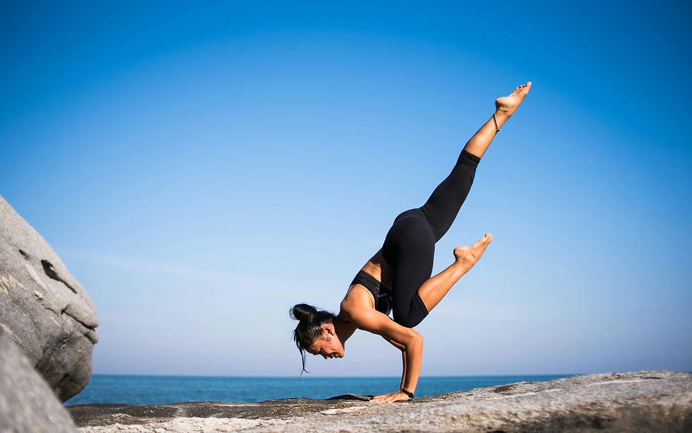 Woman practicing yoga on rock by the sea, balancing on hands with one leg extended. Clear blue sky and ocean create a serene mood.