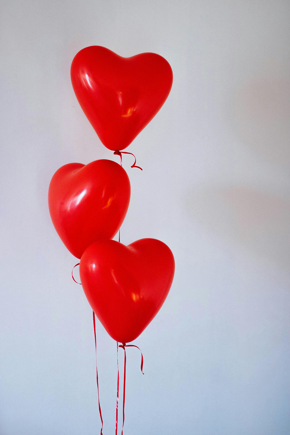 Three red heart-shaped balloons float against a plain white background, creating a cheerful and romantic atmosphere.