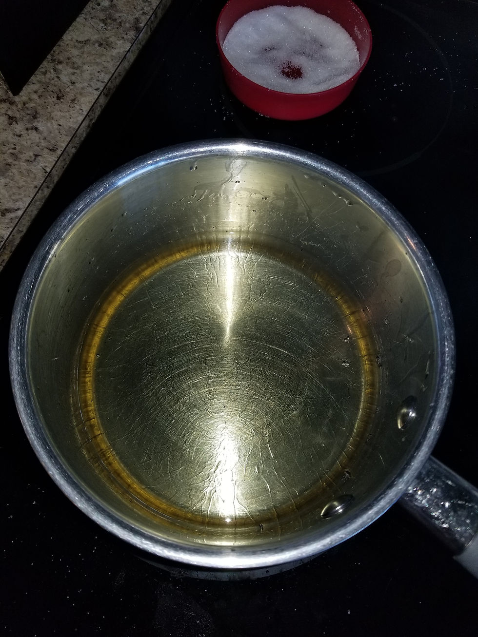 In the front, a sauce pan full of oil to fry the doughnuts. In the back, small red bowl with sugar.