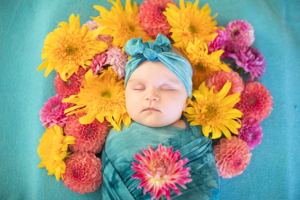 Newborn baby sleeping surrounded by yellow and pink flowers, wearing a teal headwrap