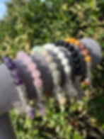Various gemstone bracelets with silver charms displayed with lush green foliage background.