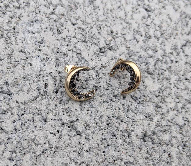 Gold crescent moon earrings with black stones displayed on textured gray surface.