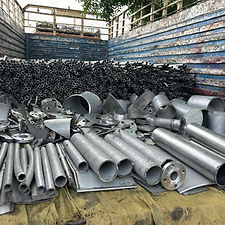 A pile of shiny, assorted metallic scrap, including pipes, sheets, and other industrial pieces, lies in the back of a truck.