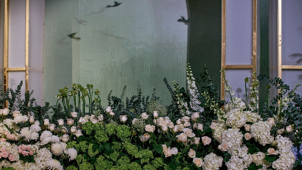 A lush arrangement of green and white flowers and foliage in front of large window panels.