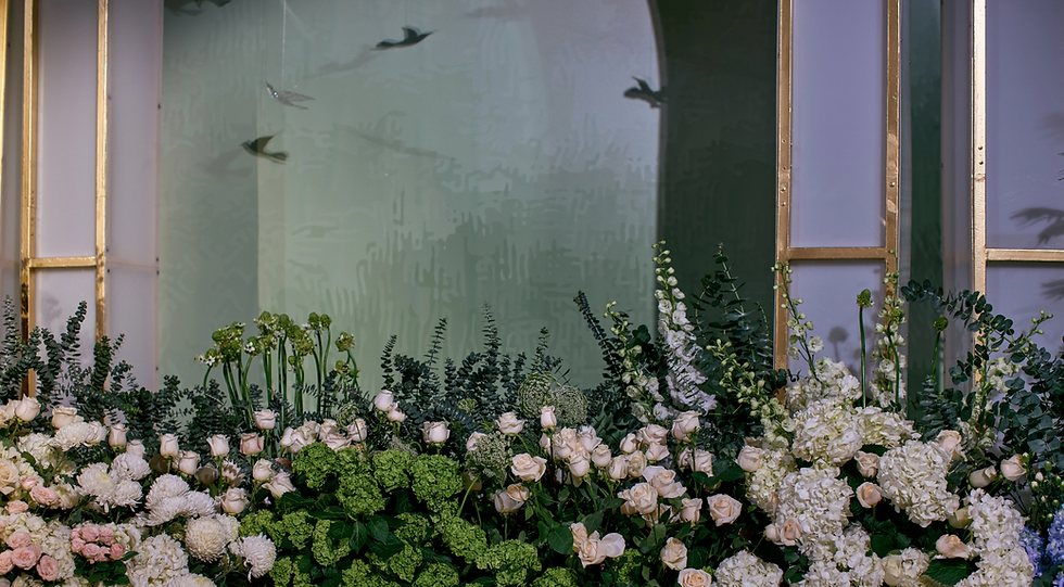 A lush arrangement of green and white flowers and foliage in front of large window panels.