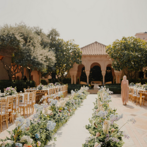An elegant outdoor wedding reception with a floral aisle leading to a building with arched architecture