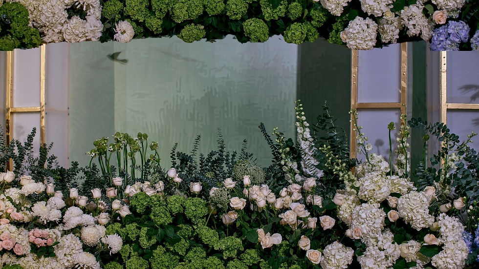 A lush arrangement of green and white flowers and foliage in front of large window panels.