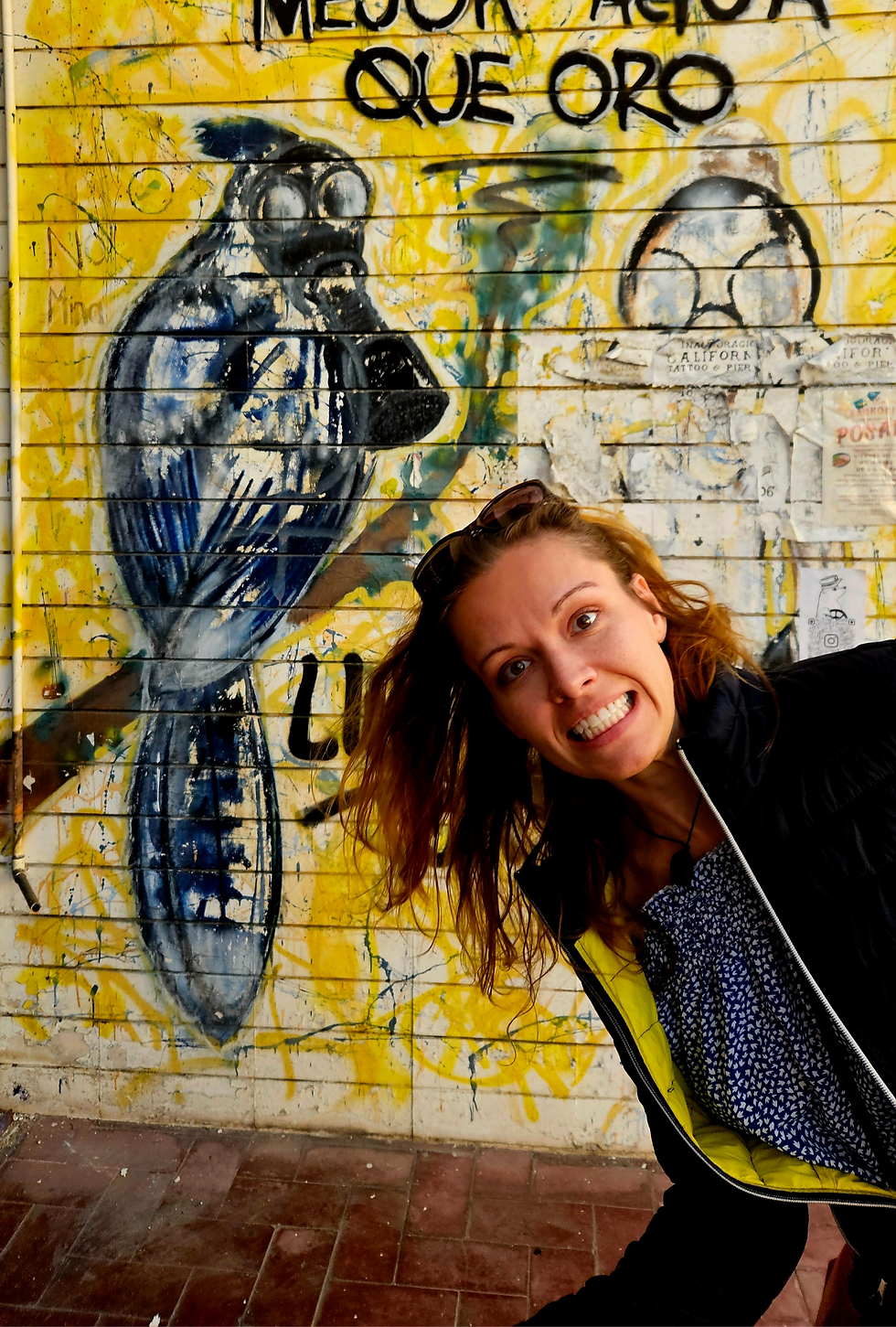 Žena s rozesmátým výrazem pózuje před barevnou pouliční malbou ptáka s plynovou maskou a nápisem ve španělštině "Mejor agua que oro" (Voda je lepší než zlato). Pozadí tvoří žlutá a modrá graffiti na staré zdi.