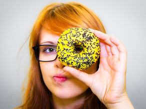 zrzavá dávka v černých brýlí držící žlutý donut s kousky čokolády před levým okem