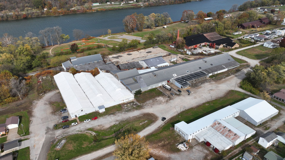 Aerial view of an industrial complex with white and gray buildings near a river, surrounded by trees with autumn foliage and a small residential area.
