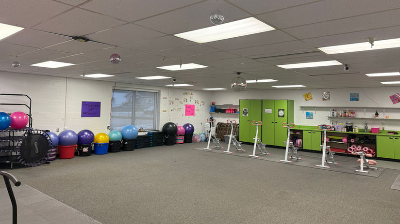 A brightly lit fitness room with colorful exercise balls, stacked mats, and stationary bikes. Green cabinets line the right wall. 