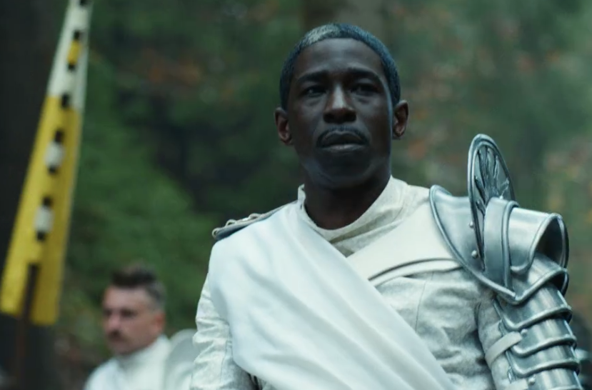 Abdul Salis as Eamon Valda wearing full Whitecloak regalia including silver pauldron on his left arm. In the background you see another Whitecloak holding the sun banner.