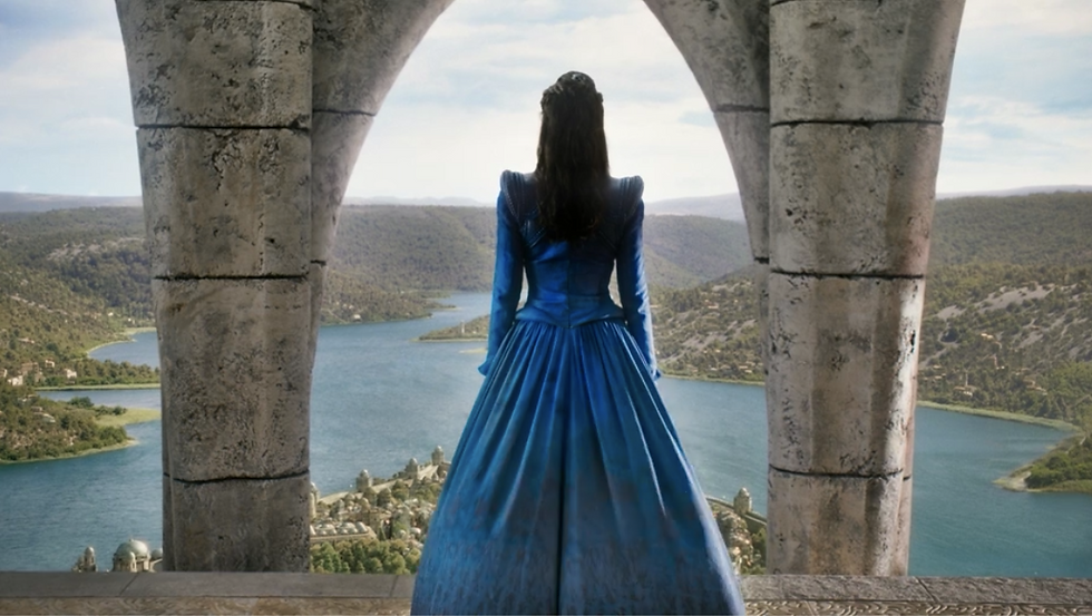 Moiraine standing at the top of the white tower, framed by columns, overlooking the harbor, wearing a long blue gown