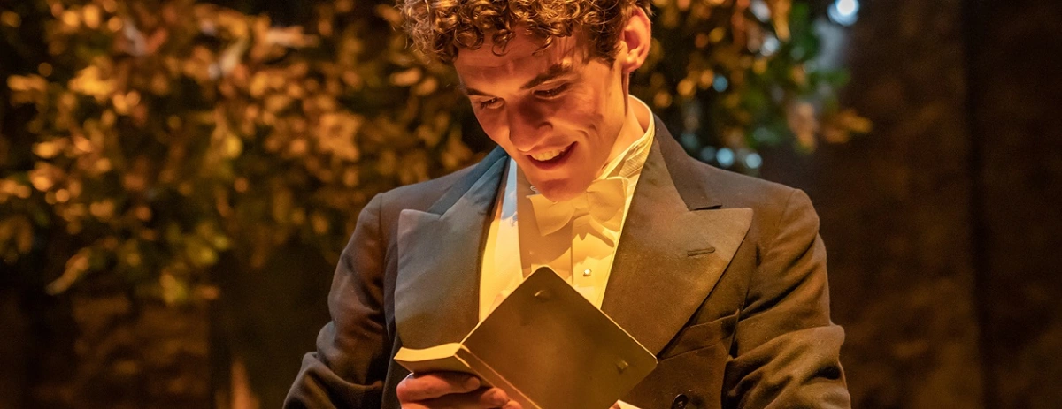 Donal Finn wearing an old fashioned tux in a warmly lit set, looking down at a small booklet and smiling. There appears to be a tree behind him.