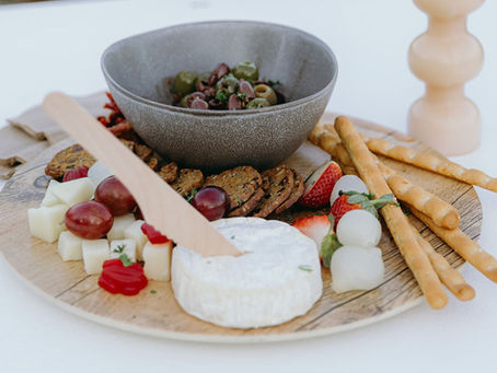 A round wooden grazing board topped with a wheel of soft cheese, fresh grapes, strawberries, cubed cheeses, olives in a bowl, breadsticks, crackers, and assorted antipasto items.