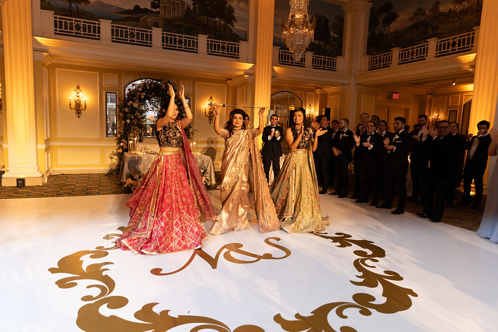 Three women in colorful dresses perform a Persian dance on a custom-decorated floor, surrounded by applauding guests in a lavish hall with chandeliers.