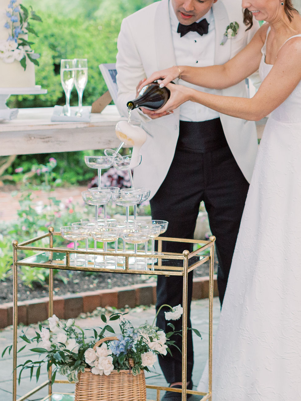 Bride and groom pour champagne into a cascading glass tower outdoors. A white cake and flowers decorate the setting, creating a festive mood.