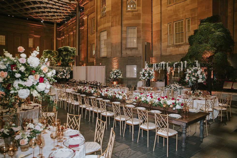 Elegant indoor wedding setup with long and round tables adorned with floral arrangements, gold chairs, and soft lighting against a grand building backdrop.
