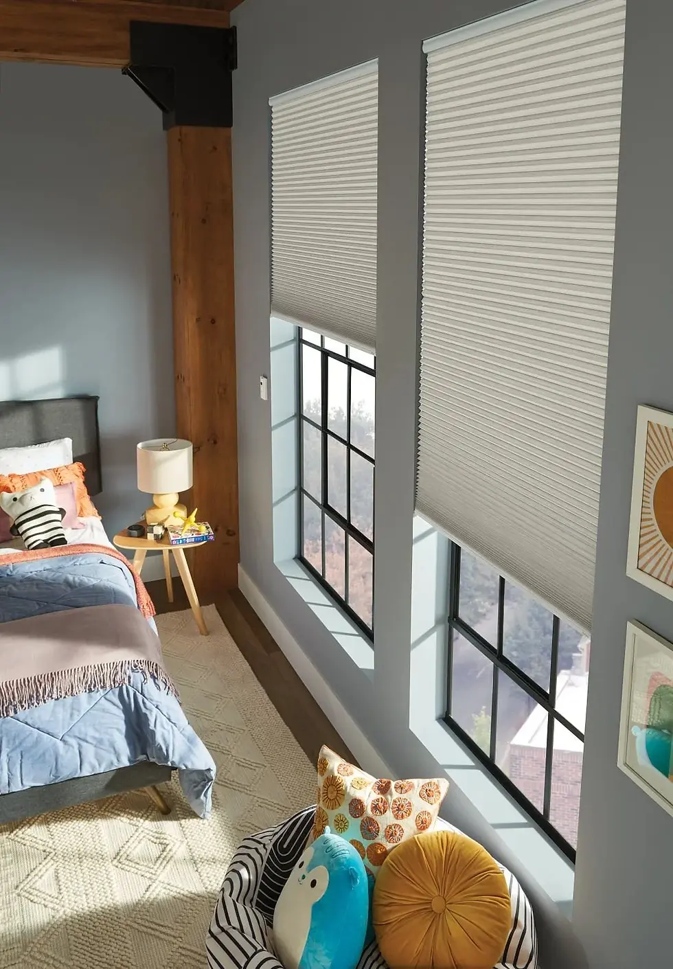 Bedroom with a cozy vibe. Blue bedding, orange throw, and plush toys on the bed. Two large windows with cellular shades. Soft morning light.