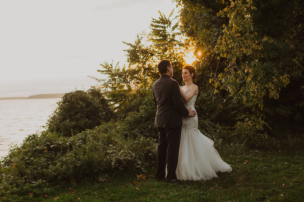 Bride and groom embrace in a lush garden by a lake at sunset. Bride in white dress, groom in dark suit, radiant golden sunlight.