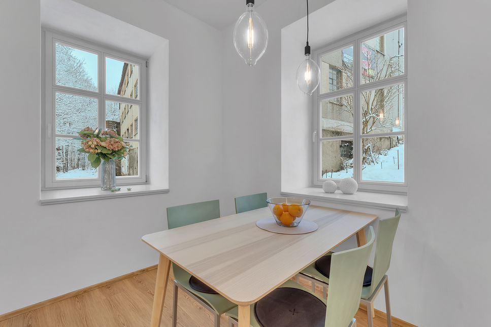 Na obrázku je jídelna spojená s obývacím pokojem. Stůl je umístěn mezi dvěma okny, takže je velice světlá. Stůl je z jasanové dýhy a židle jsou v mátově zelené barvě. Nad stolem je osvětlení, které tvoří dvě skleněné baňky. Místnost je vymalovaná bíle a doplňuje jí podlaha v imitaci dubu.