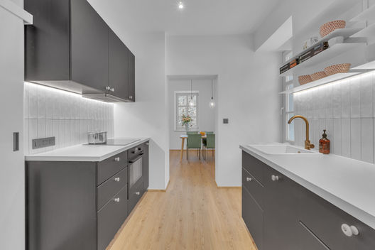 Na obrázku je kuchyn, s protilehlými pracovními plochami. Má černou nebo antracitovou barv, bílé keramické úchytky. Je to moderní kuchyn s prvky retra, které jí dávají doplňky v barvě růžového zlata.