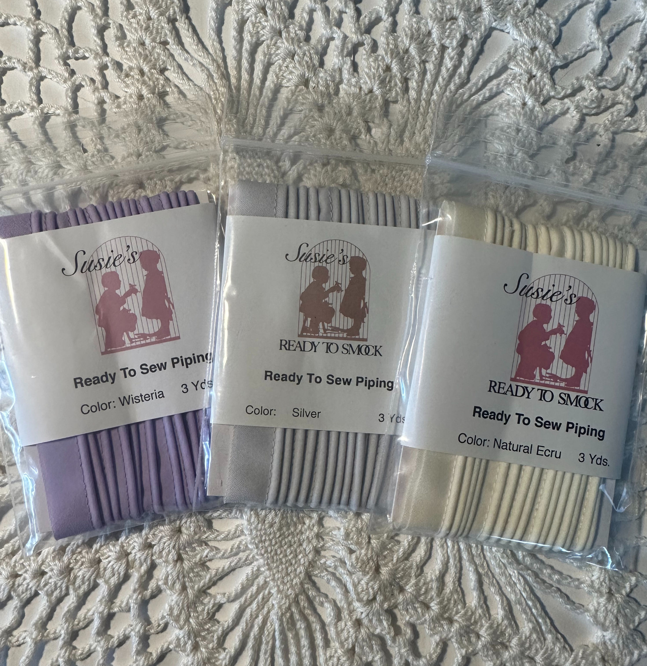 Wisteria, Silver and Natural Ecru sewing trim packages on a lacy cloth