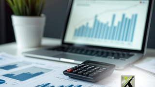 A calculator and printed financial charts on a desk with a laptop displaying a bar graph in the background, representing tax preparation and business analysis. The Xpert Business & Tax Solutions logo is in the corner.