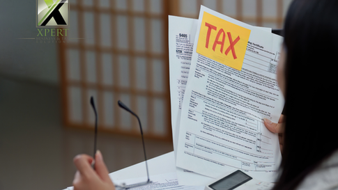 Person reviewing tax documents with a yellow note labelled “TAX,” holding paperwork at a desk with a calculator, representing tax preparation and financial review, branded with the Xpert Business & Tax Solutions logo.