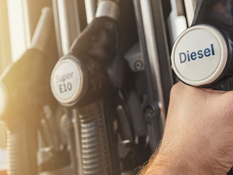 Hand holding a diesel fuel nozzle at a gas station, with Super E10 and Diesel pump labels in warm sunlight.