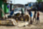 Men and boys gather around wire cages and a white car on a dusty street, with a green Postes sign and market shacks.