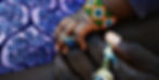 Close-up of a baby's hand holding an adult's finger, with a colorful beaded bracelet over purple patterned fabric.