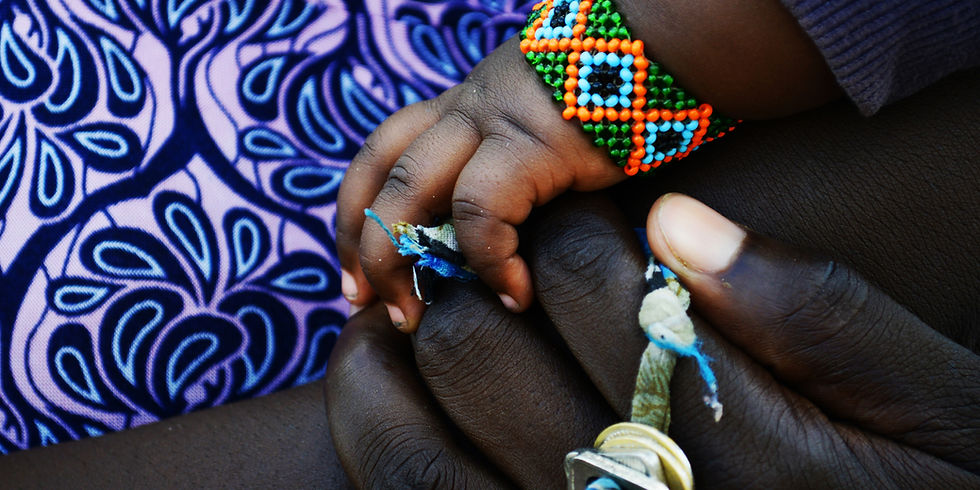 Close-up of a baby's hand holding an adult's finger, with a colorful beaded bracelet over purple patterned fabric.