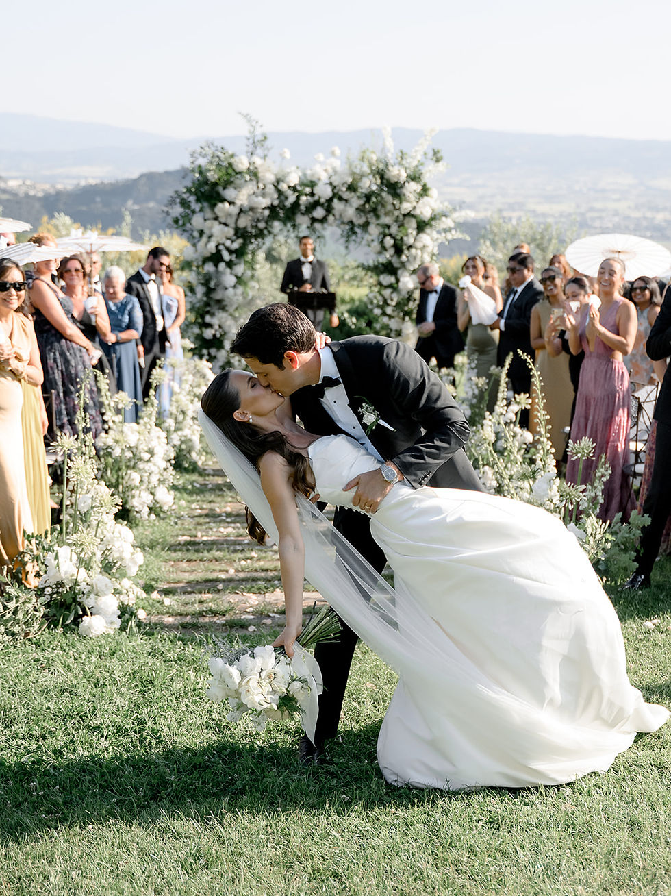 Bride and groom sharing their first kiss during an outdoor destination wedding ceremony in Italy with a white floral arch and panoramic Umbrian hills view