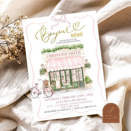 Baby shower invitation with a pink shop illustration, floral accents, and gold text. Details include date, time, and location in elegant script.