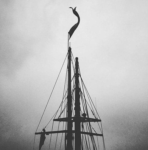Black and white ominous image of the rigging of a ship with flag blowing in a harsh wind