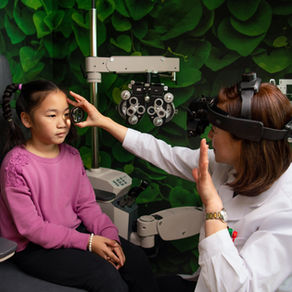 Eye doctor examines a young girl’s eye with a lens in a clinic, with green leaf-pattern wallpaper behind them.