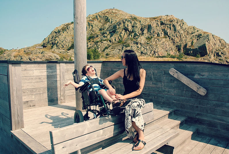 A woman with long black hair sits with a boy in a wheelchair wearing sunglasses