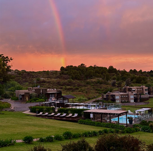 Complejo turístico con piscinas y áreas verdes bajo un cielo rosado al atardecer, con un arcoíris formando sobre las colinas del fondo.