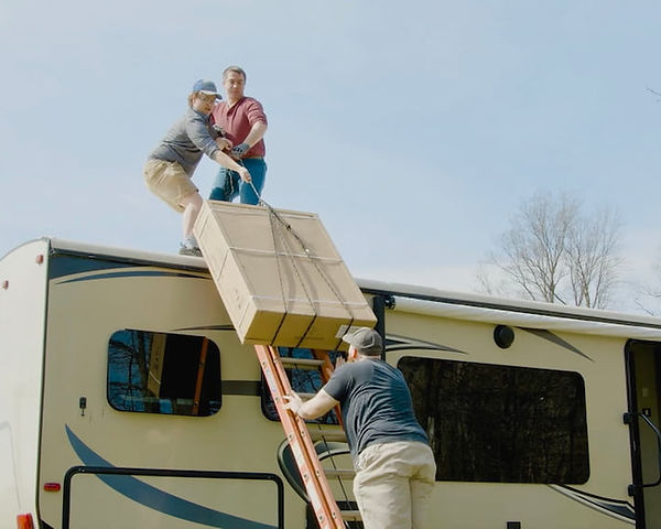 3 people working together to slide an air conditioner box to the top of an RV roof.