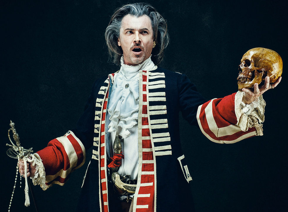 Man in 18th-century attire holds a skull, gesturing dramatically. Dark background, red and black coat, ruffled shirt; theatrical mood.