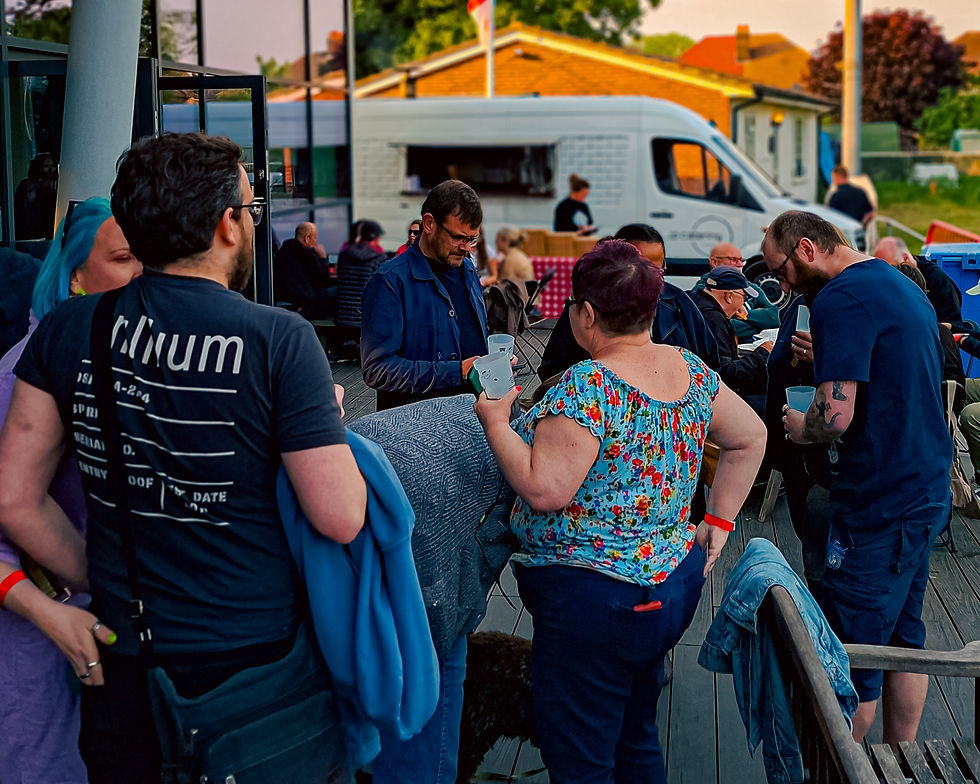 A group of people socializing outdoors near a food truck. A person in a floral top holds a drink. Background shows picnic tables and trees.