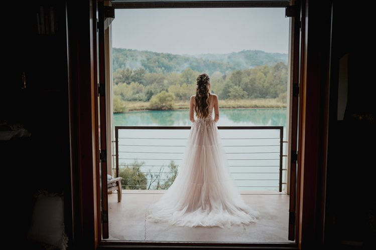 Bride on balcony overlooks lake.