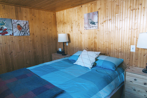 A queen sized bedroom with a blue duvet over, two pine nightstands with lamps and pine walls.
