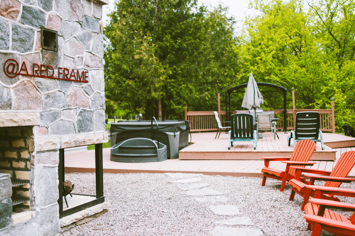 A picture of the granite fireplace, flagstone pavers lead you to the deck and into the hottub. There are four red muskoka chairs surrounding the fireplace. A expansive brown deck is perfect for entertaining or using the propane bbq.