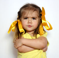 Toddler in a yellow shirt with yellow satin ribbons in her hair against a gray background. Her arms are crossed with a look of determination.