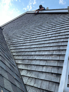 Man on steep cedar shingle roof performing maintenance under blue sky.