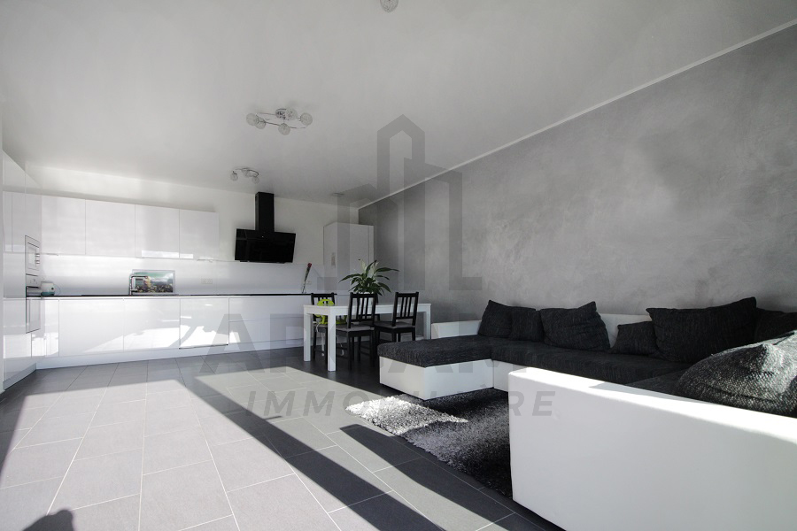Open-plan living room with a black-and-white sectional sofa, dark rug, white dining furniture, and the fitted kitchen behind.